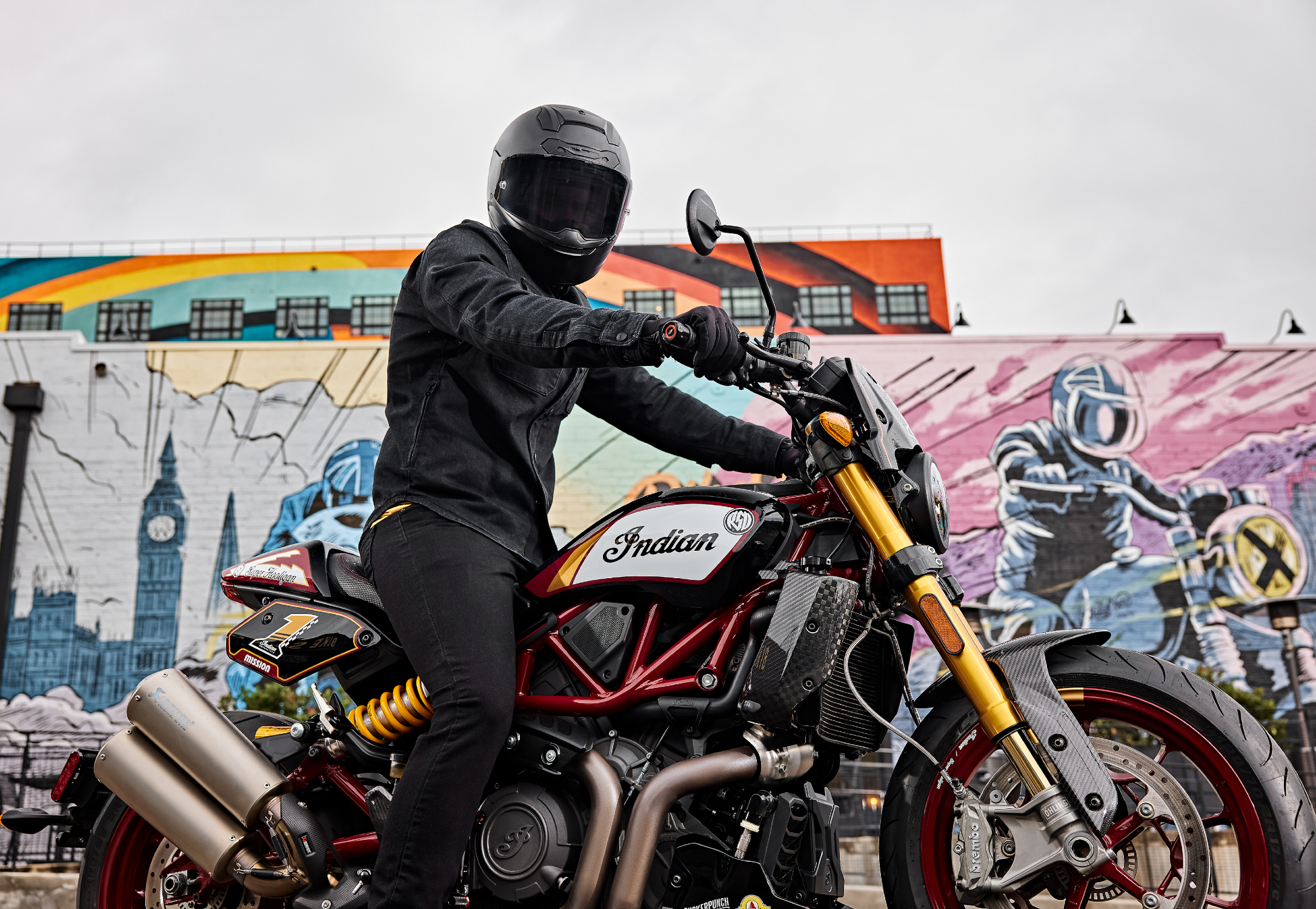 Rider wearing all black moto gear sitting on 2024 Indian FTR x RSD Super Hooligan in front of graffiti art Bike Shed wall.
