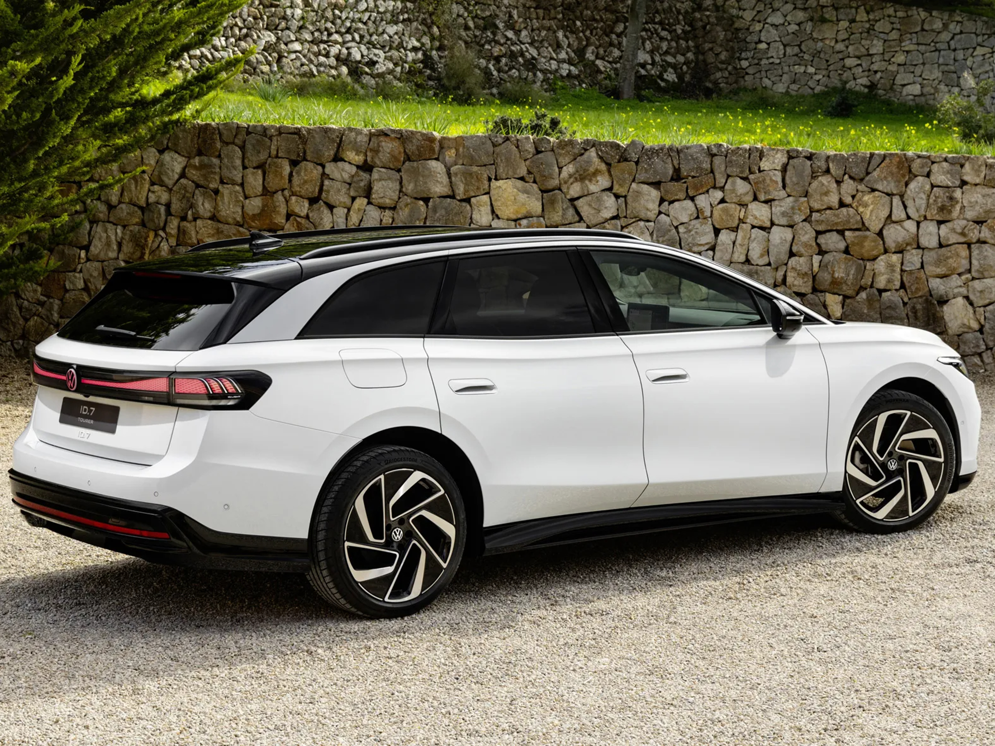 VW ID.7 concept vehicle parked on a stone driveway with a landscaped rock wall in the background right rear three-quarter view of white car.