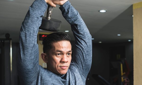 Man in gym wearing long sleeve blue shirt doing overhead tricep extension exercise with weight