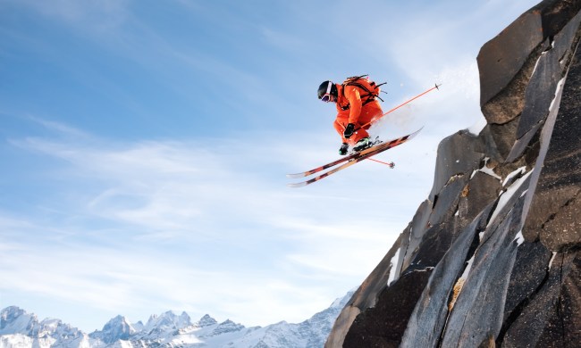 A professional skier makes a jump-drop from a high cliff against the blue sky leaving a trail of snow powder in the mountains. Caucasian ridge in the background. Photo from the slopes of Mount Elbrus. The concept of extreme sports and recreation in the mountains in winter. Copy the space