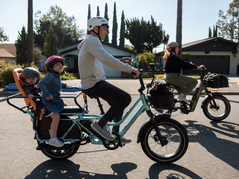 Two adults riding on Rad Power Bikes e-bkes, the adult in the foreground is riding a RadWagon 5 with two kids in child seats on the back rack.