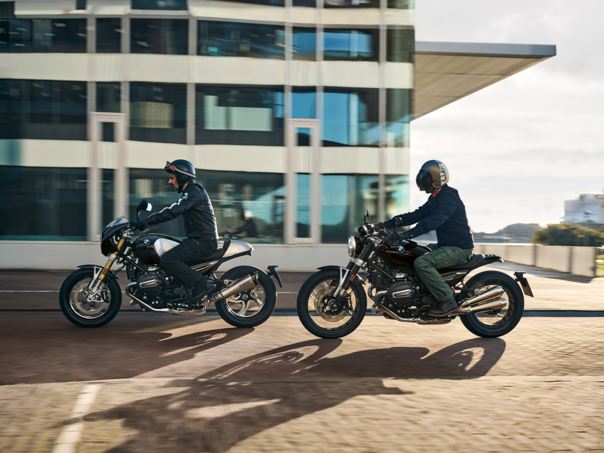 Two riders on 2024 BMW R12 and R12 NineT motorcycles driving from right to left on a brick road in front of an office building.