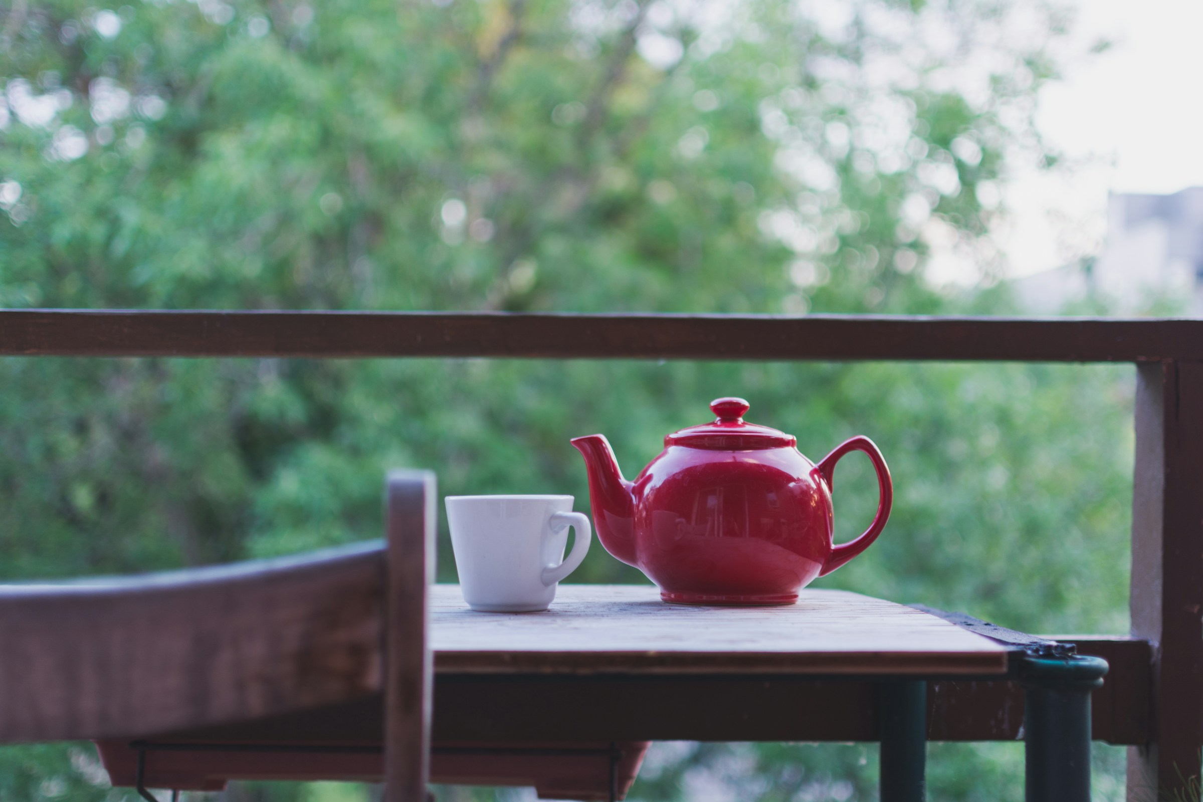 red teapot and white cup on wooden table outside with green trees in background