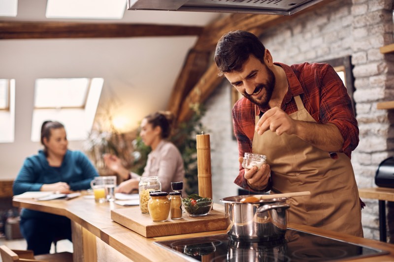 Happy man adding salt while cooking for his friends in the kitchen.