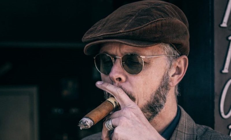 Man smoking a cigar in a newsboy cap outside.