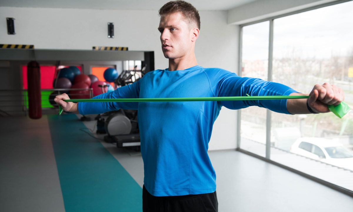 man wearing blue shirt stretching with resistance band performing band pull apart exercise in gym