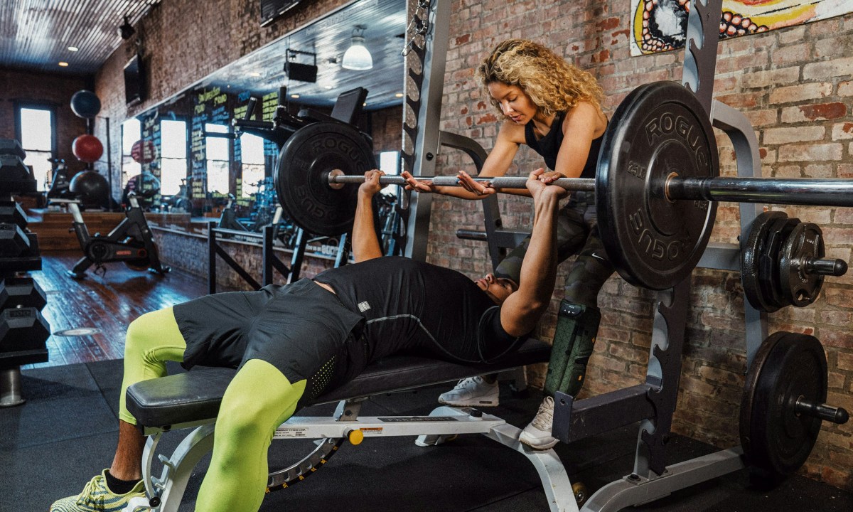 Man bench pressing in gym wearing green pants on a bench woman helping train him