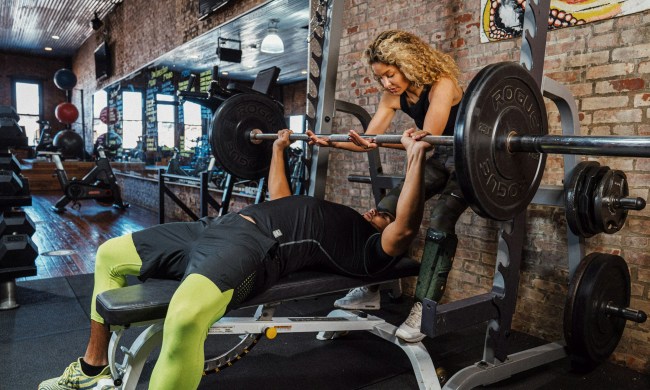Man bench pressing in gym wearing green pants on a bench woman helping train him