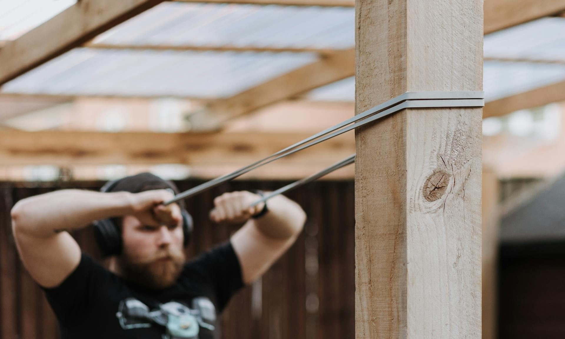 Man wearing black T shirt cap and headphones doing face pulls with resistance bands on a wooden pole