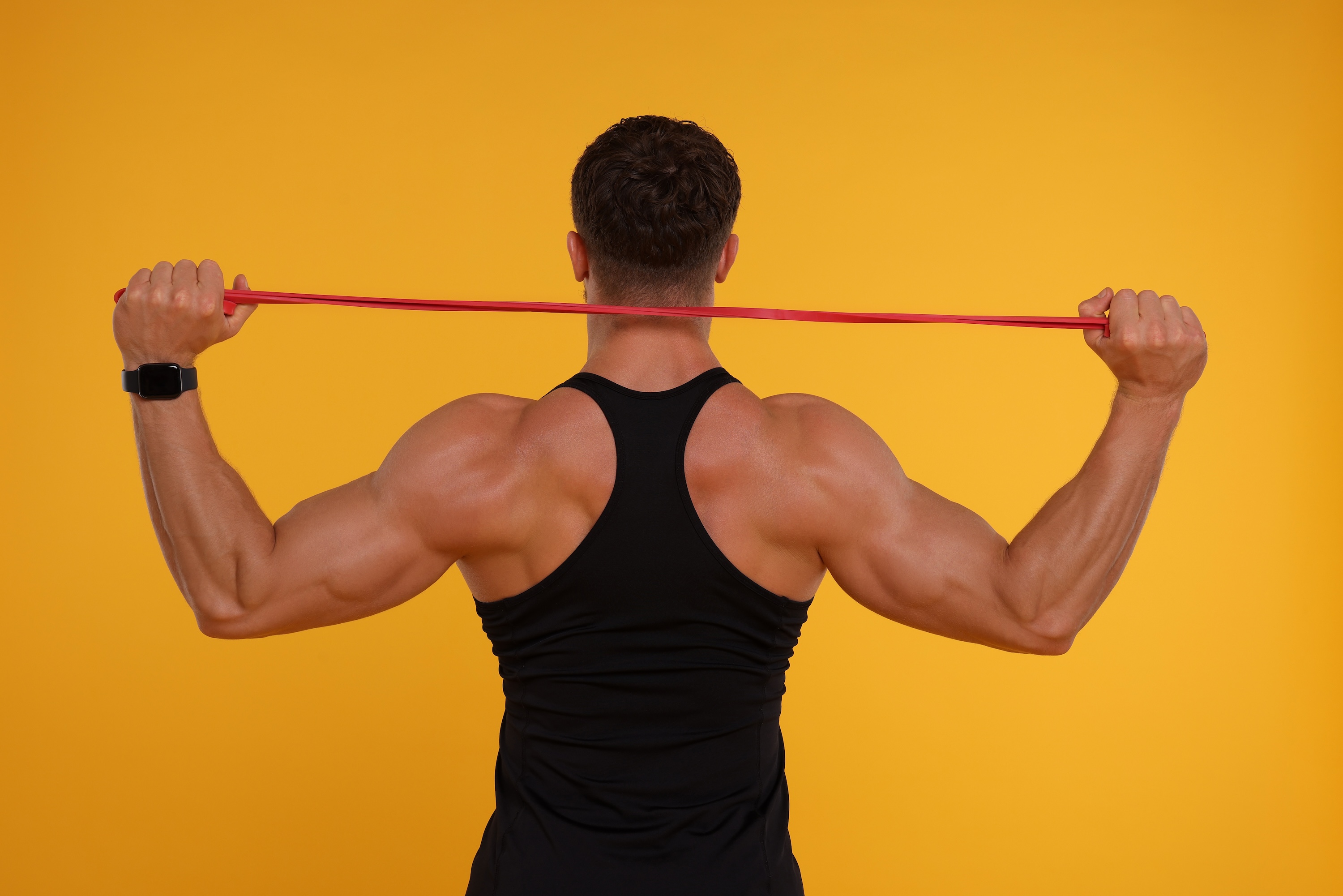 Young man exercising with elastic resistance band on orange background, back view