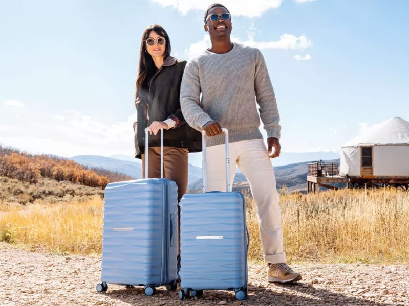 A couple stand in nature with Samsonite suitcases.