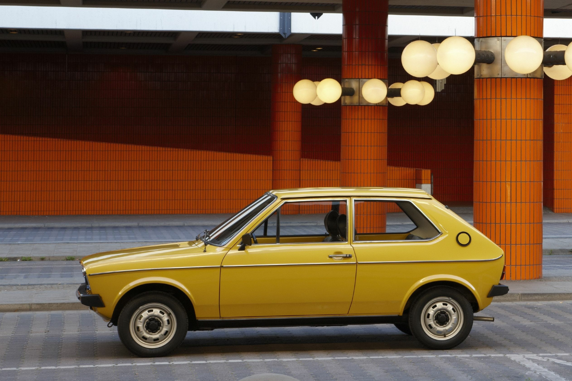 A yellow Audi 50 pared on the side of a roadway with a large red concrete building in the background.