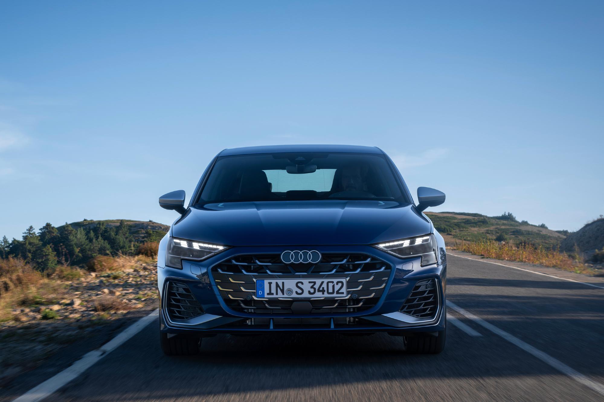 Blue 2025 Audi S3 direct front view with the car driving on a mountain road with rocks and trees on either side.
