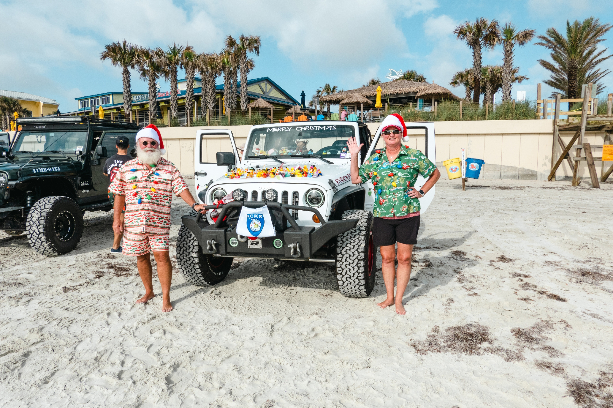 Photo from Jeep Beach Week 2023 with Santa and Ms. Claus on the beach with their white Jeep Wrangler.
