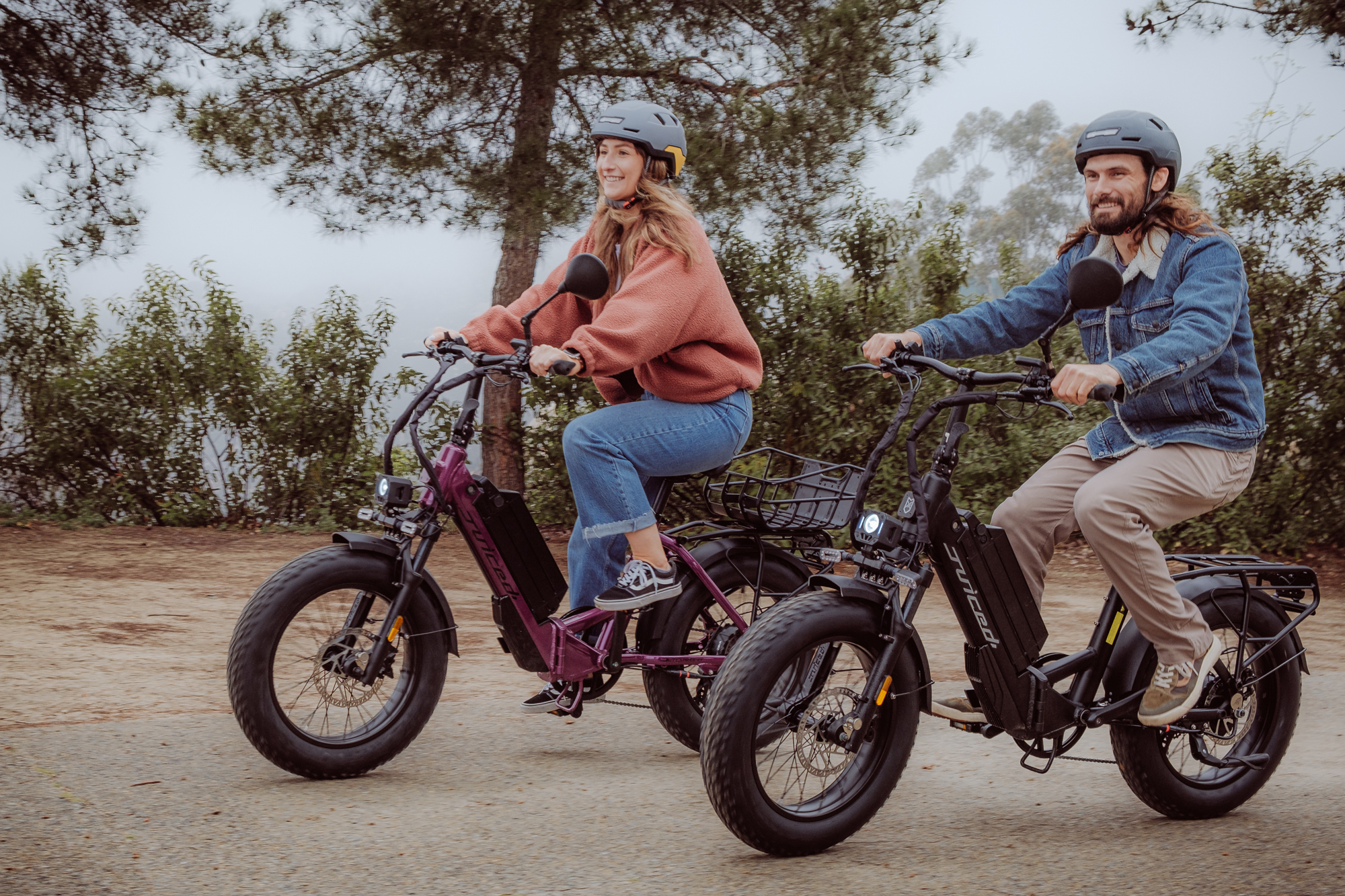 Two people riding Juiced Bikes JetCurrent Pro e-bikes on a forest path, Purple Haze and Black colors.