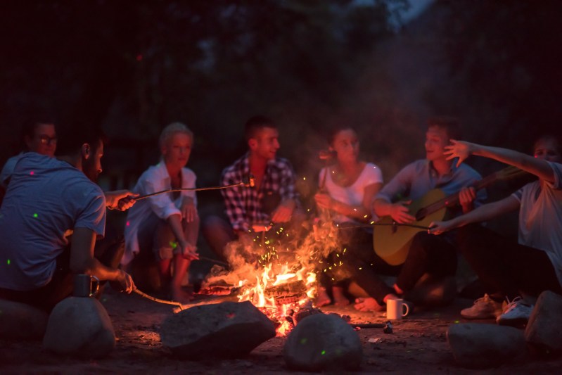 a group of happy young friends relaxing and enjoying summer evening around campfire on the river bank