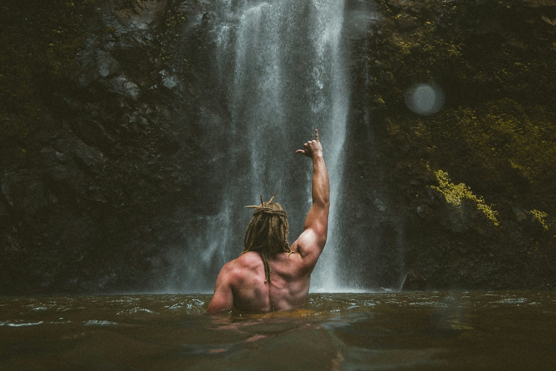 shirtless man lifting arm up making fist outside in water by waterfall nature muscle man