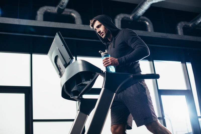 low angle view of handsome sportsman in hoodie exercising on treadmill and holding sport bottle with water in gym