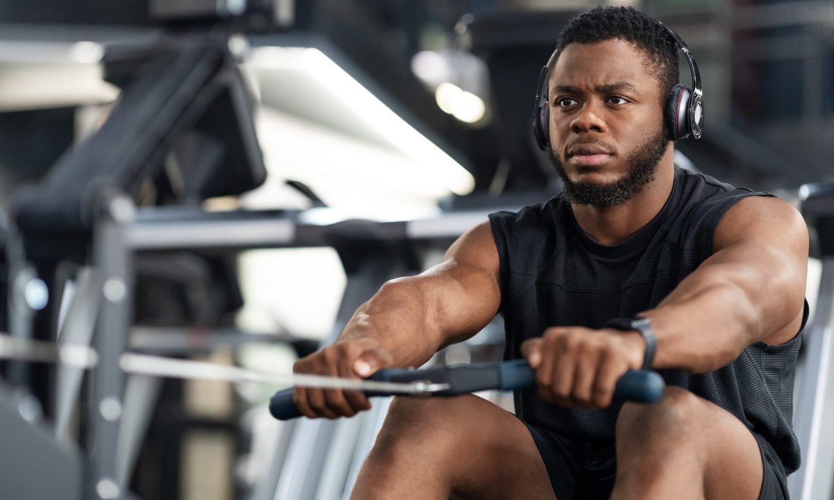 Closeup of handsome african american sportsman training body on modern rowing exerciser in gym, copy space. Motivated young black sportsman with wireless headset having workout on rowing machine