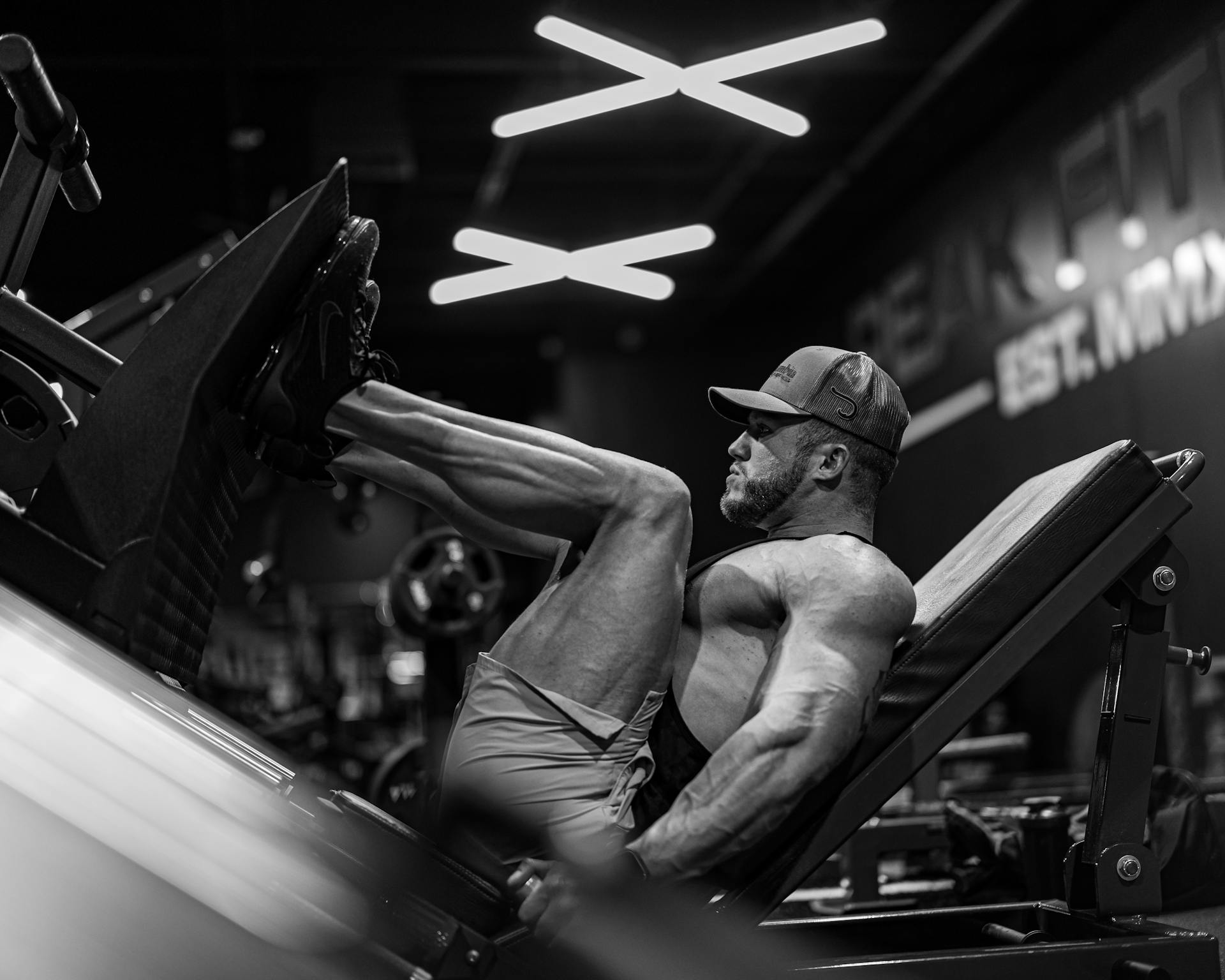 black and white image of man in gym wearing hat and shorts performing leg press exericse on machine