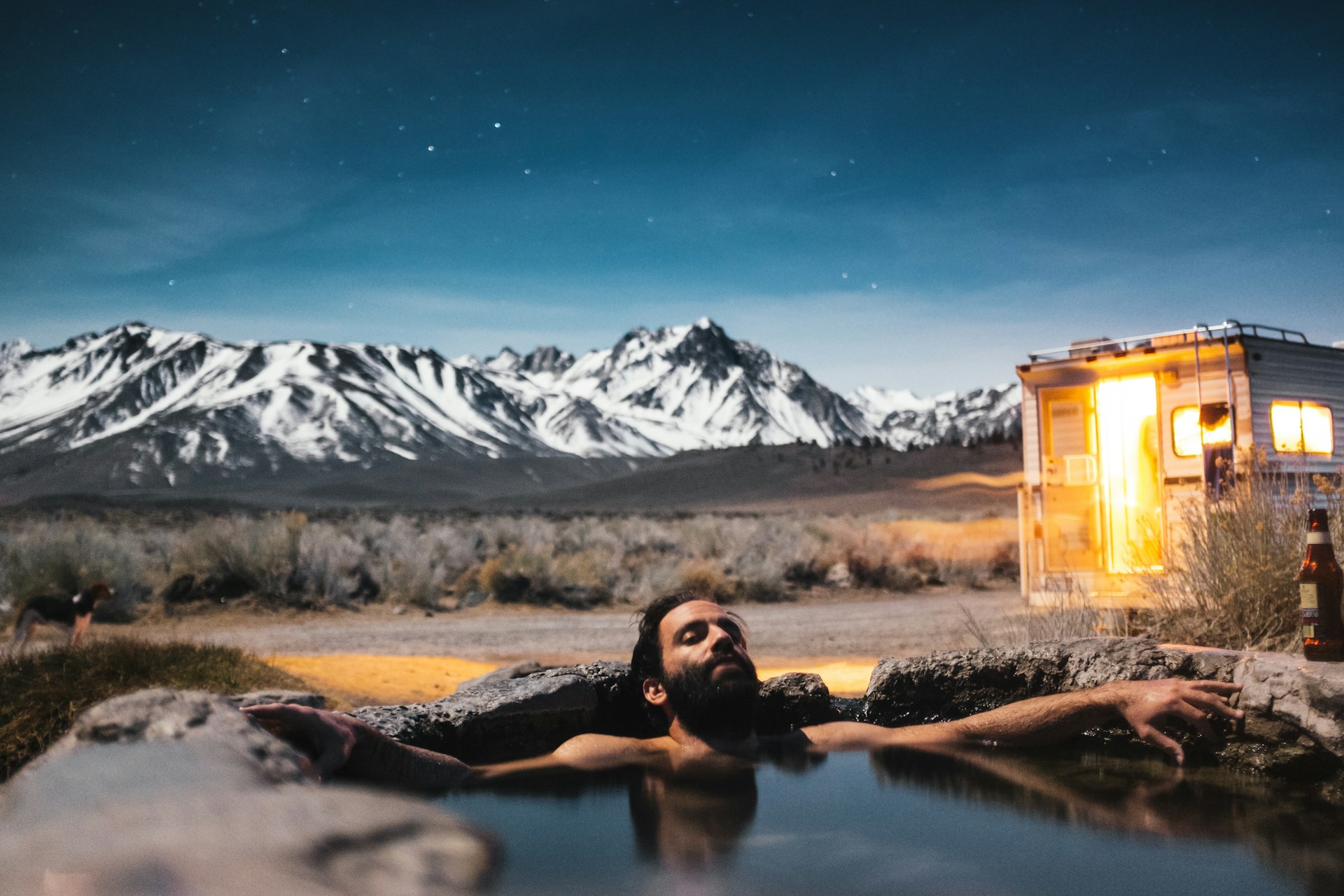 A man soaking in a natural tub outside of an RV during dusk with a mountain range behind him.