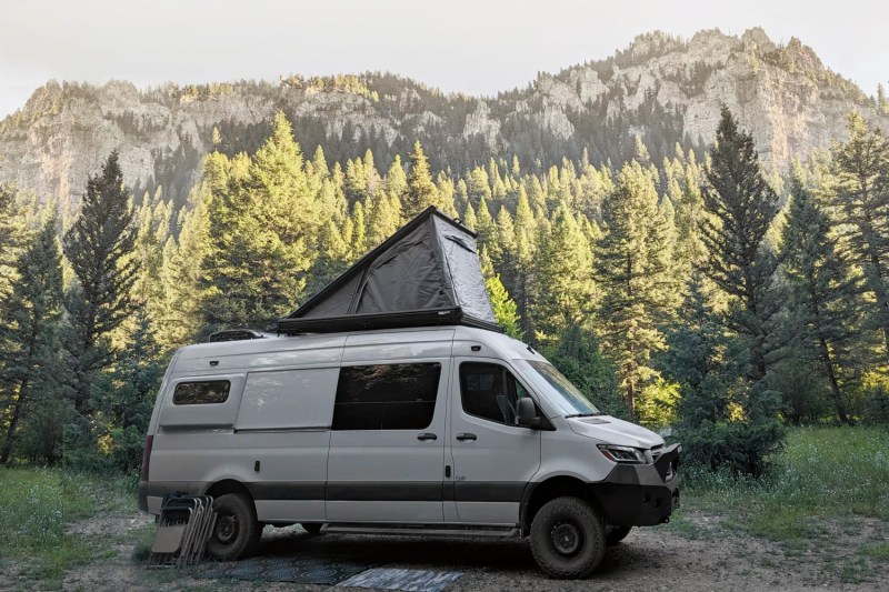 Camper van outfitted with Super Pacific's CloudCap pop-up roof tent parked among a stand of trees.