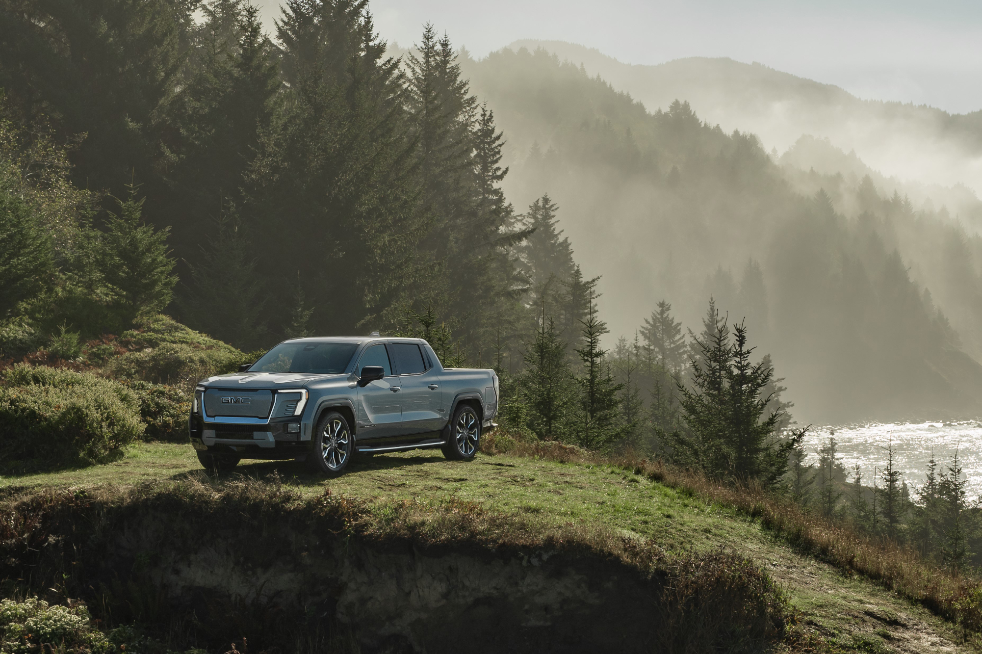 2024 GMC Sierra EV Denali Edition 1 parked on a grassy hill in an area with trees, mountains, fog, and rough ocean coastal water.