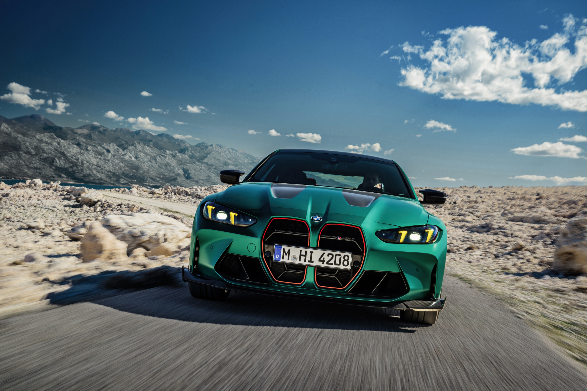 2025 BMW M4 CS driver direct frontal shot driving on single-lane blacktop in a rocky desert setting with mountains in the background.