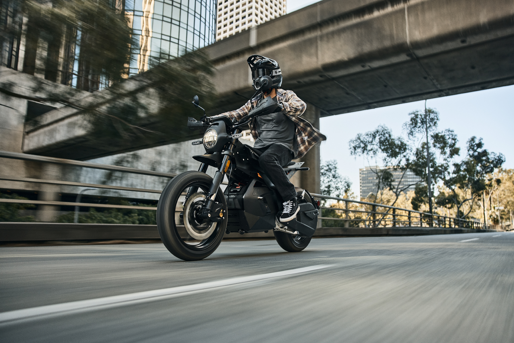 A rider on a Ryvid Outset electric motorcycle in Vapor Grey on a city street under an overpass.