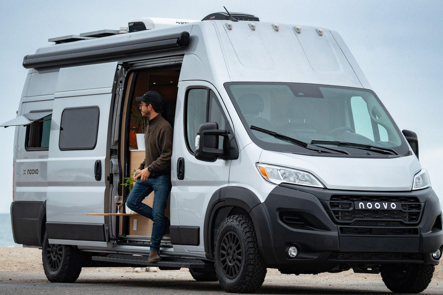 Man stepping out of a Noovo Plus camper van parked near the beach.