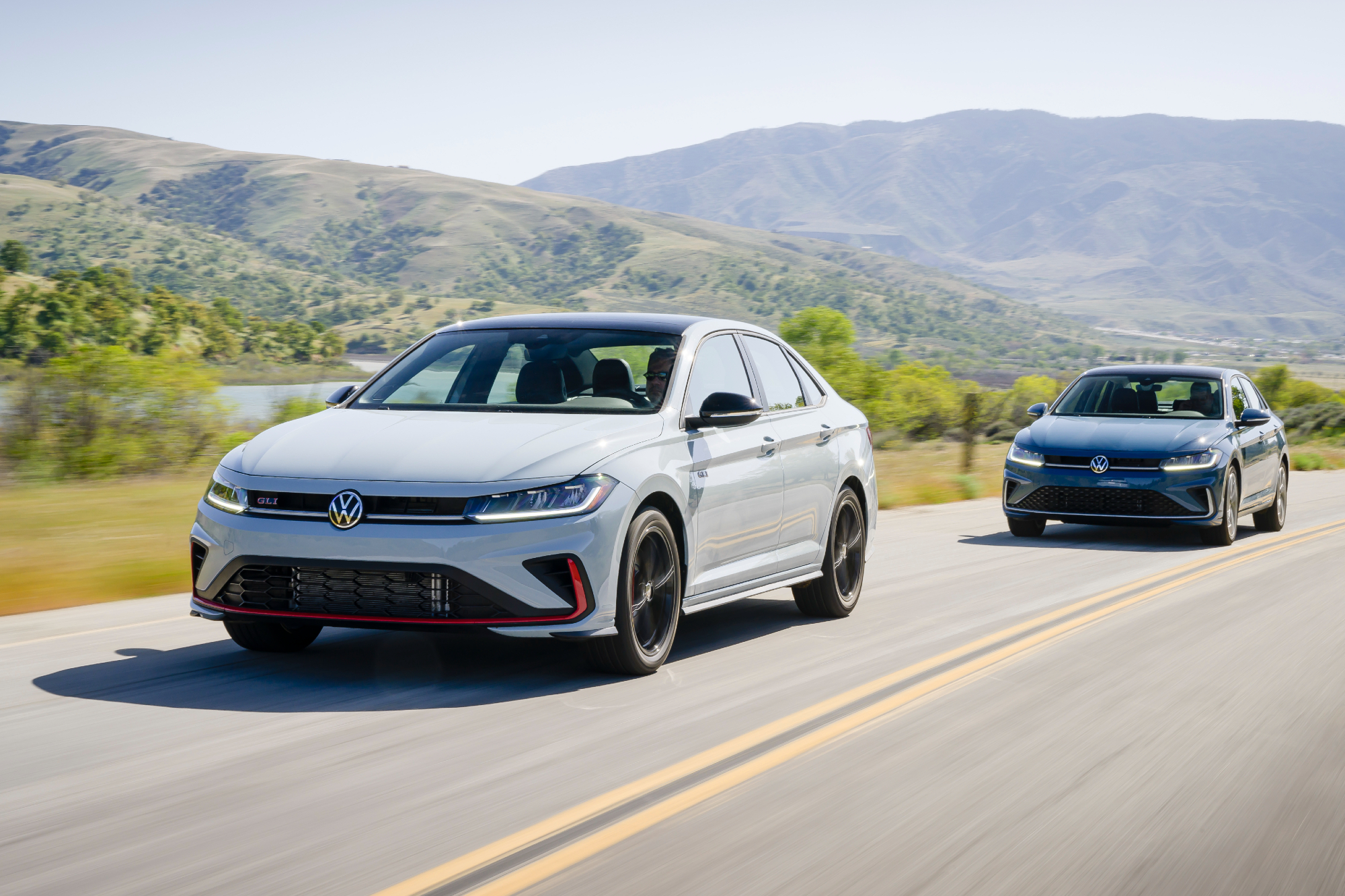 2025 VW Jetta GLI and 2025 VW Jetta left front three-quarter image driving on paved road with mountains in the background.