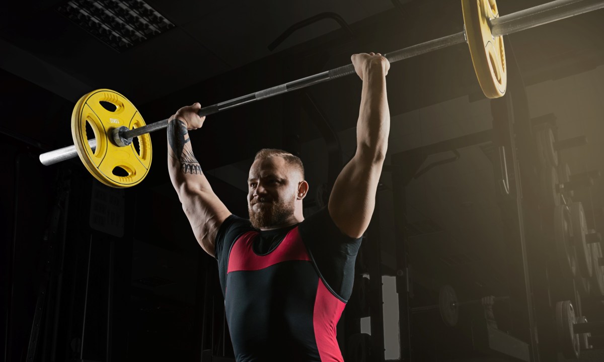 Man lifting overhead press military press exercise with barbell in gym wearing black and red shorts