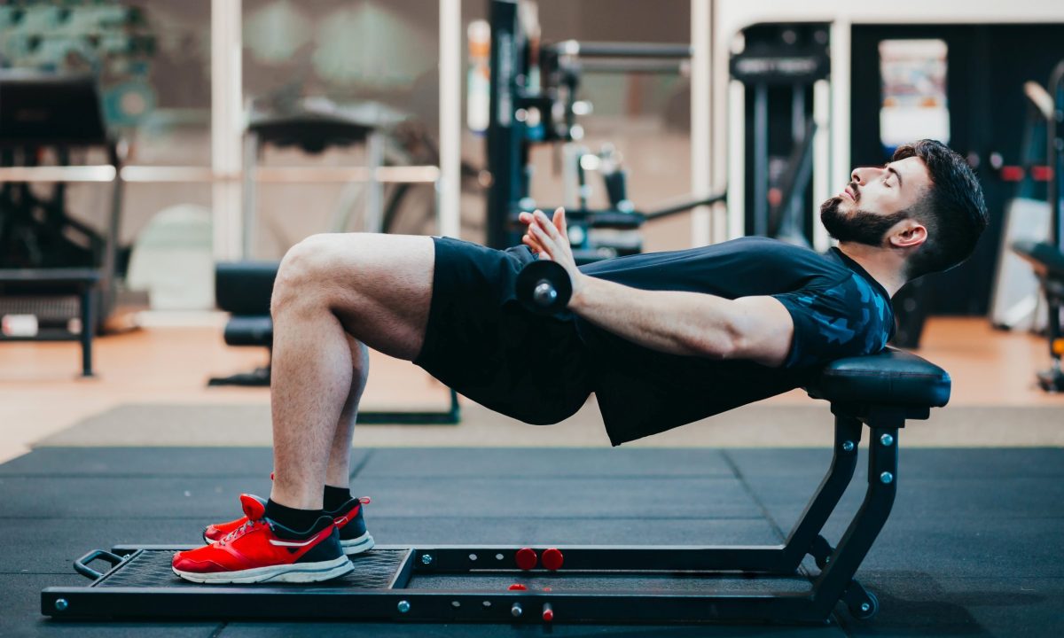 Man wearing black and red sneakers in gym doing hip thrusts with weight barbell on bench