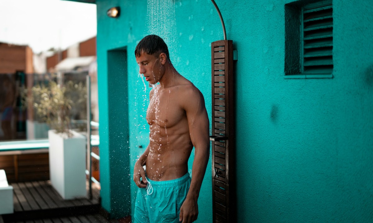 Shirtless man outside in blue shorts showering by blue wall