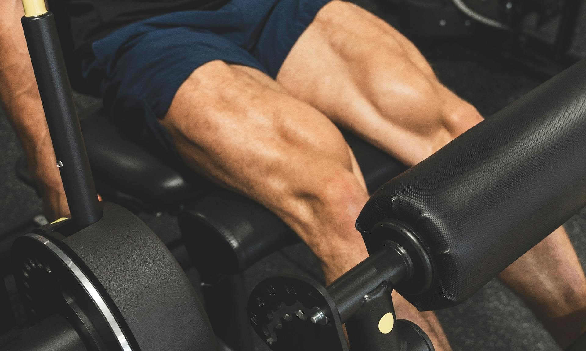 Man wearing black shirt and blue shorts close up doing leg machine seated leg curl in gym
