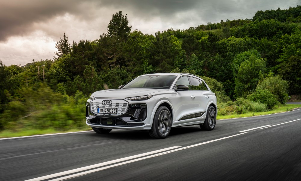 White Audi Q6 e-tron drivng on highway with forested hills in the background left front three-quarter view.