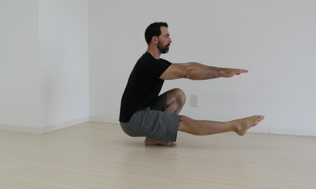 Man in a black T-shirt doing pistol squat inside on wooden floor