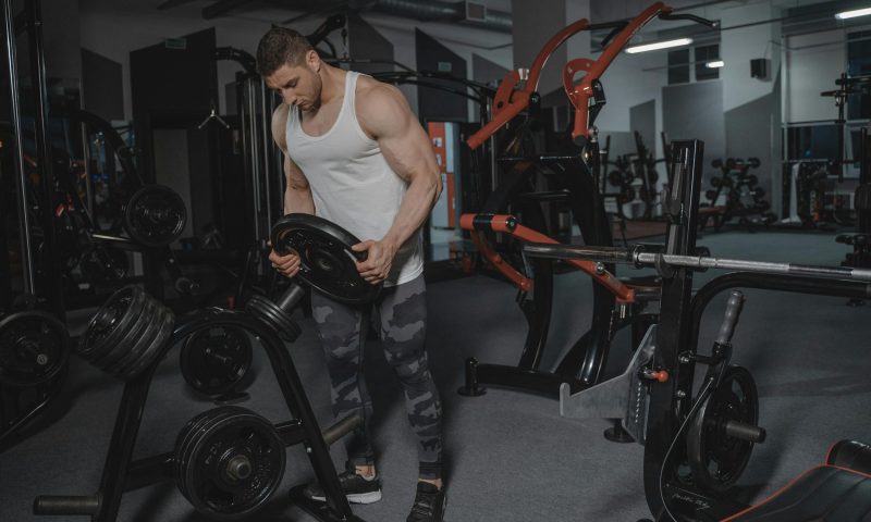Man wearing a white tank top in the gym holding a weight and stacking weights on a rack