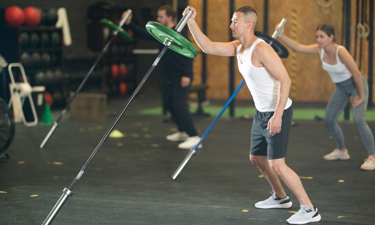 Man and people in gym doing standing one-arm landmine press exercise with barbell landmine attachment