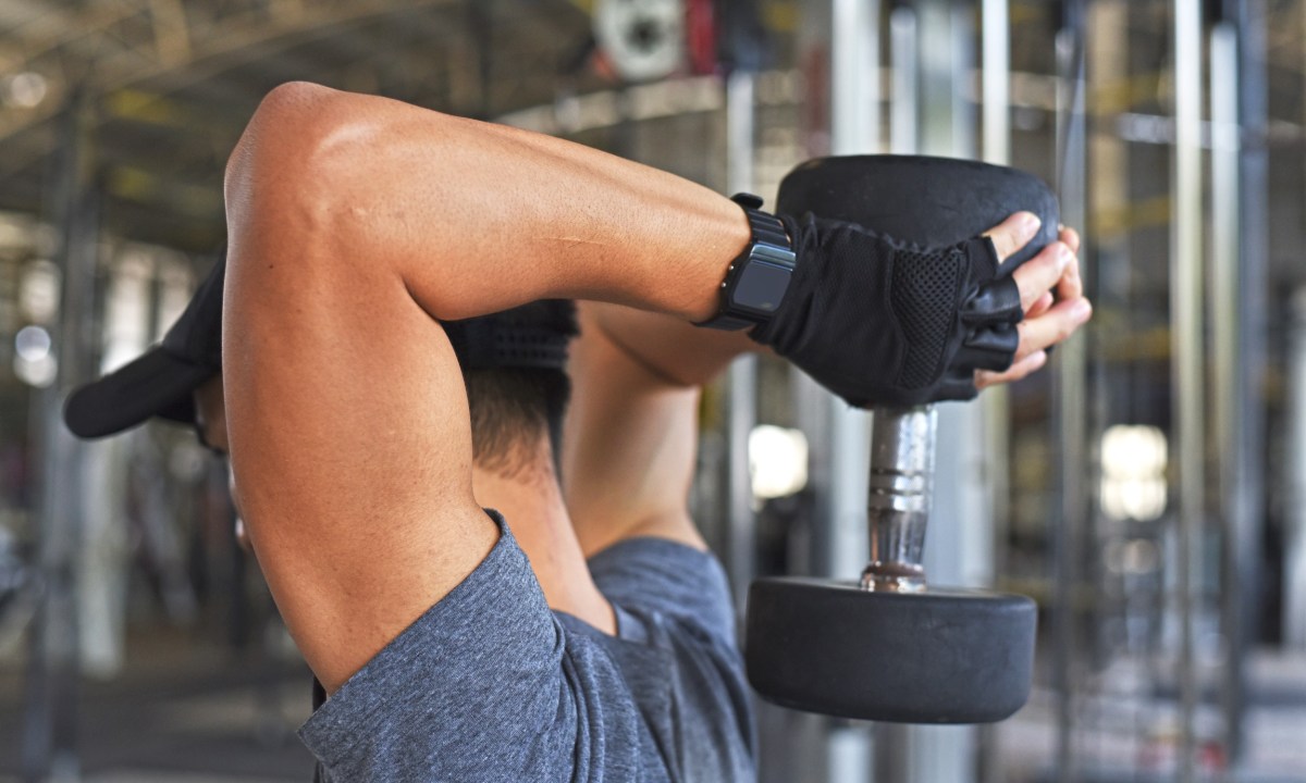 Man in black hat and blue T-shirt doing an overhead tricep extension with dumbbell in the gym