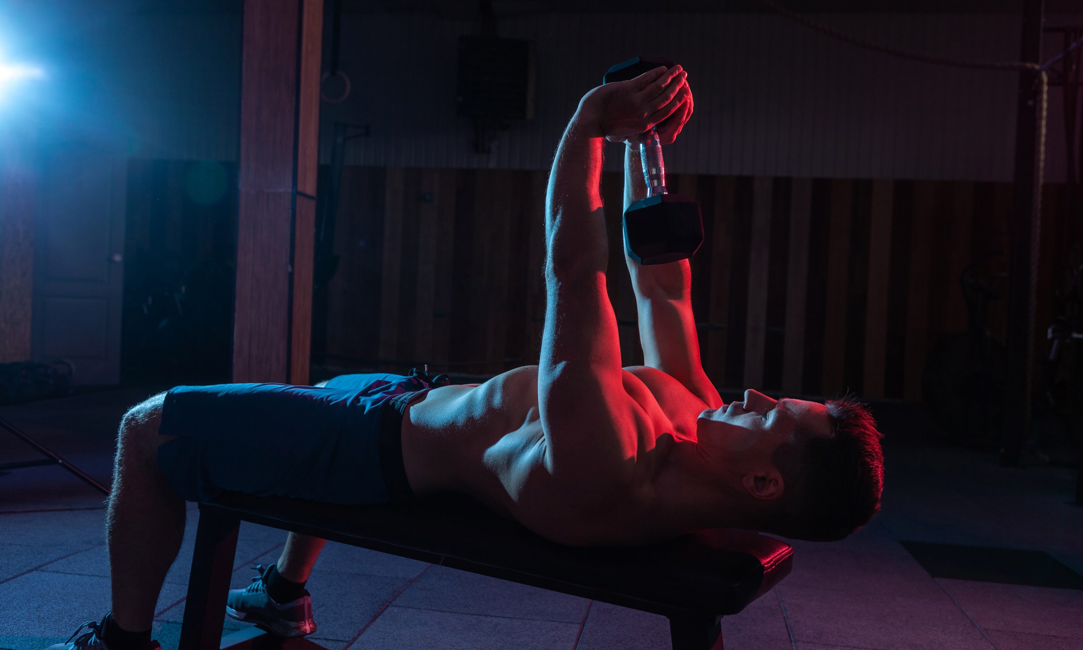Shirtless man on bench doing dumbbell pullover in red light