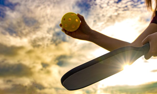 Woman holding a Pickleball paddle and ball as she prepares to serve with the sun rising behind her.