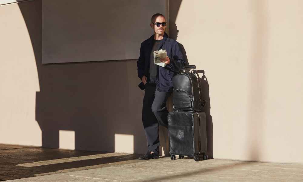 man standing by wall with a suitcase and a bag