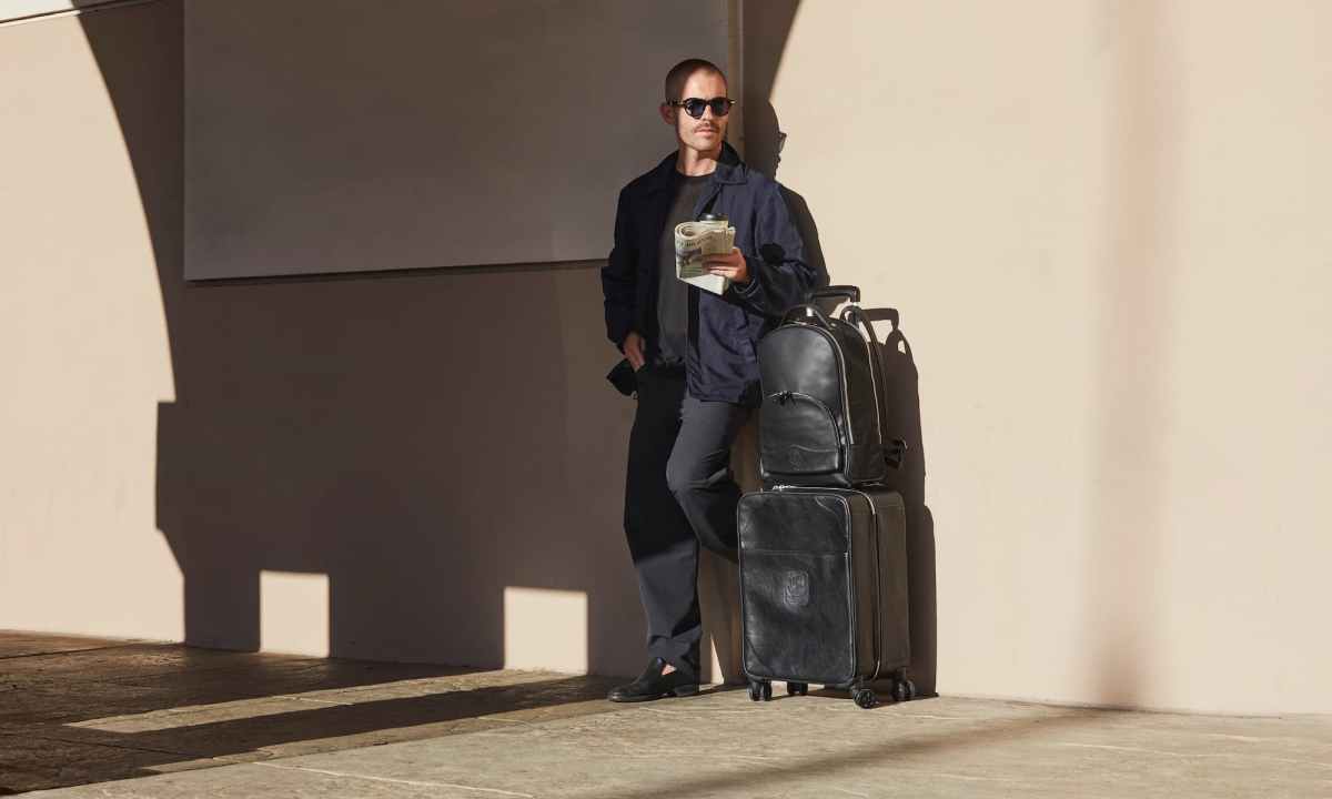man standing by wall with a suitcase and a bag