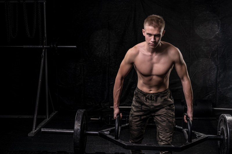 Shirtless man on a dark background performing trap bar deadlift with hex bar