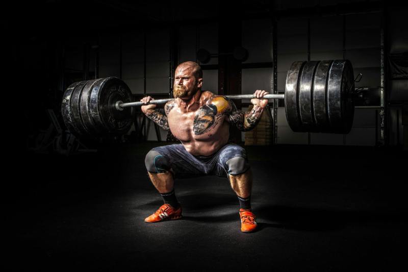 man performing barbell back squat shirtless tattoo dark backround heavy weight