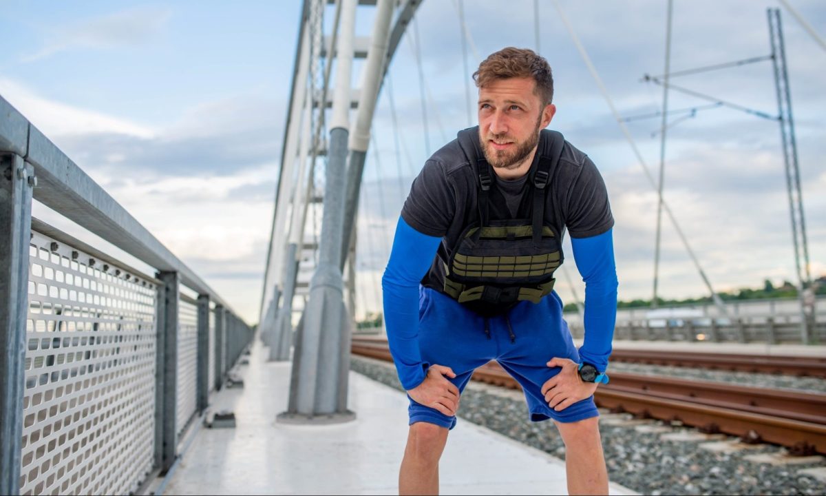 Man taking break from running working out walking wearing weighted vest outdoors in city near train track
