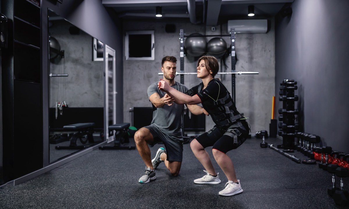 man helping woman do squats wearing EMS suit exercise electrical muscle stimulation