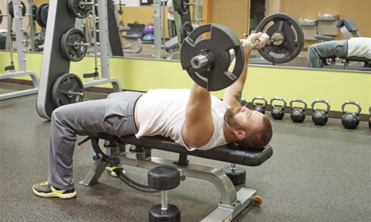 Man doing skull crushers with an EZ curl barbell on bench gym