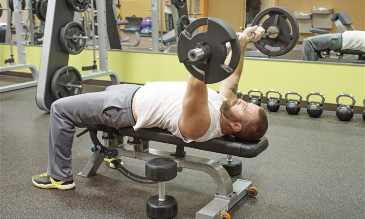 Man doing skull crushers with an EZ curl barbell on bench gym